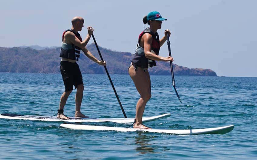 Ein Paar, das an einem sonnigen Morgen am Strand eine Tour mit dem Paddle Board genießt. 