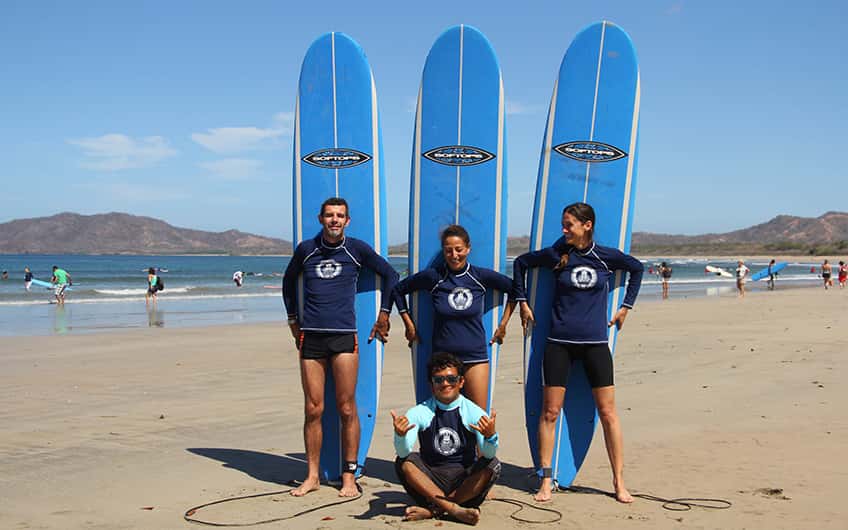 Drei Personen mit Longboards, die mit ihrem Surflehrer posieren, bevor sie Surfunterricht nehmen.