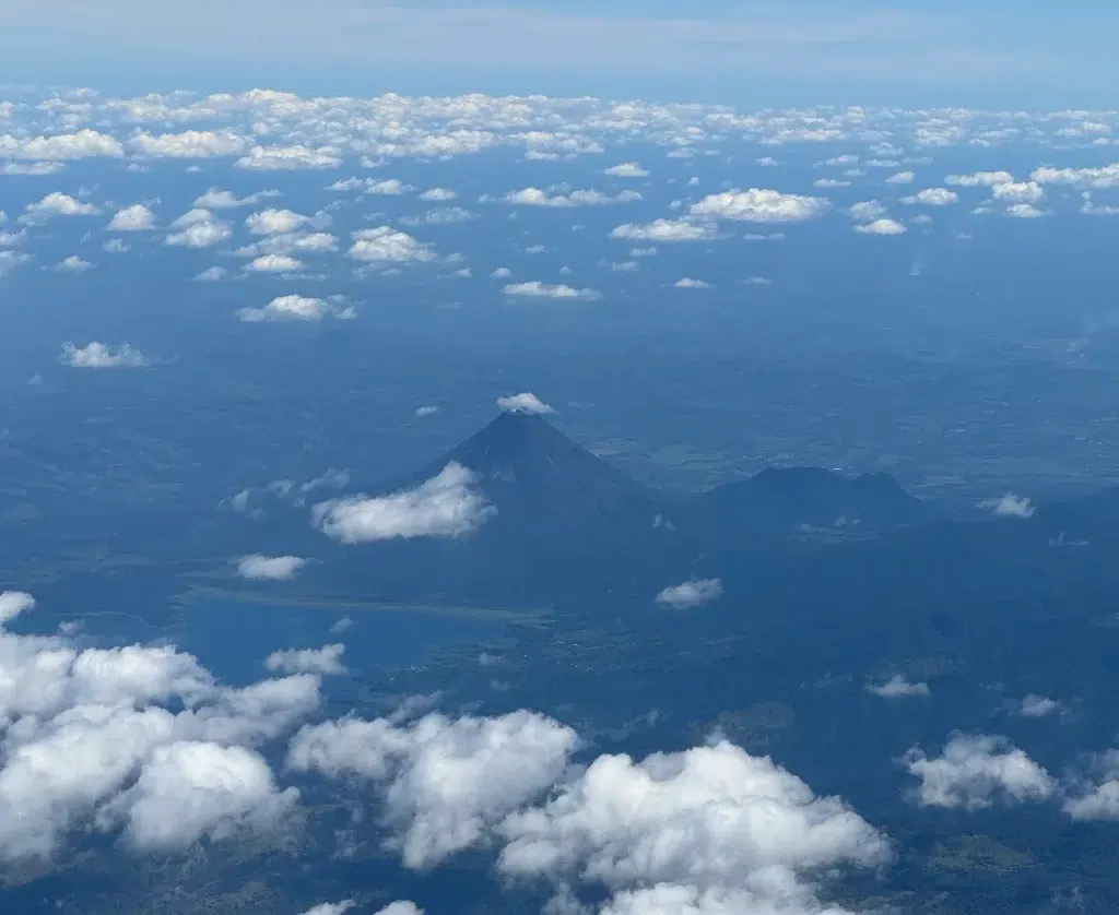 Aerial view of Arenal Volcano and Lake in Costa Rica, with the prominent conical volcano rising above surrounding landscapes and a reflective lake at its base, amidst scattered white clouds against a blue sky.